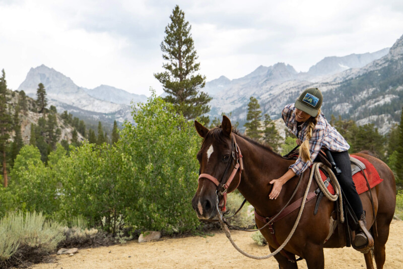 A person wearing a plaid shirt and baseball cap sits on a brown horse, patting its neck. They are outdoors surrounded by lush green trees and shrubs, with a mountain range in the background under a mildly cloudy sky in Bishop, California. visit bishop