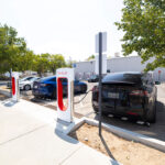 A Tesla electric vehicle is parked at a Tesla Supercharger station outside a building in Bishop, California. The charging stalls are under a clear sky and surrounded by trees. Other cars are also parked nearby, and there are signs indicating the charging spots. visit bishop