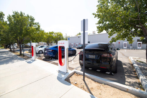 A Tesla electric vehicle is parked at a Tesla Supercharger station outside a building in Bishop, California. The charging stalls are under a clear sky and surrounded by trees. Other cars are also parked nearby, and there are signs indicating the charging spots. visit bishop