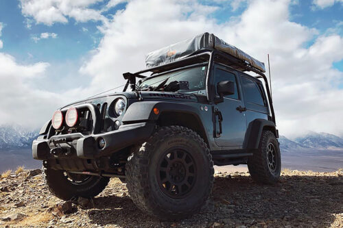 A rugged off-road vehicle with roof storage is parked on rocky terrain with mountains in the background. visit bishop