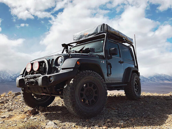 A rugged off-road vehicle with roof storage is parked on rocky terrain with mountains in the background. visit bishop