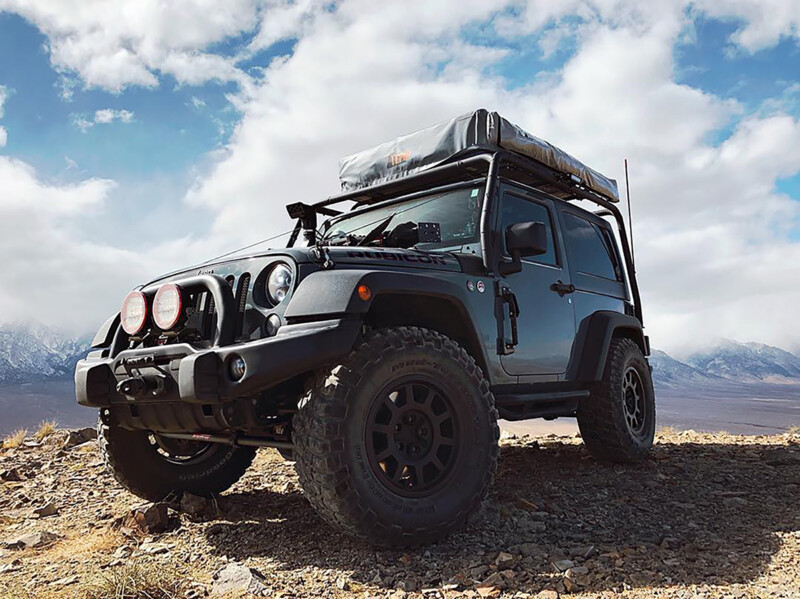 A rugged off-road vehicle with roof storage is parked on rocky terrain with mountains in the background. visit bishop