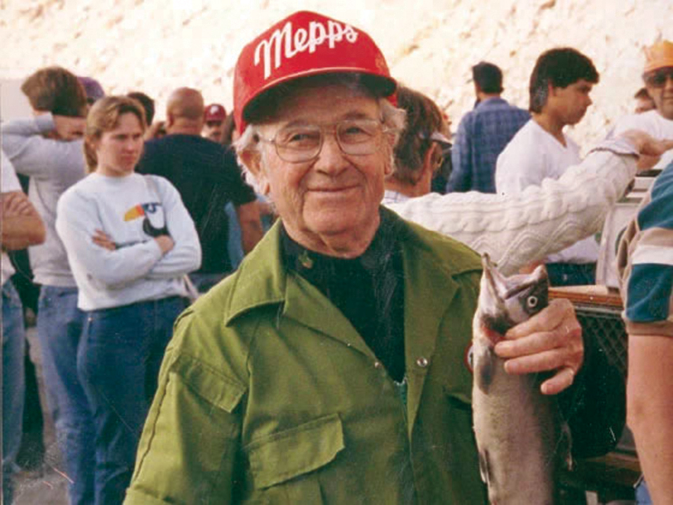 An older man wearing glasses, a green jacket, and a red cap with the word "Mepps" on it smiles at the camera in Bishop, California. He is holding a fish in his right hand. People are gathered behind him, and one person in the background is wearing a sweatshirt with a penguin graphic on it.
 visit bishop