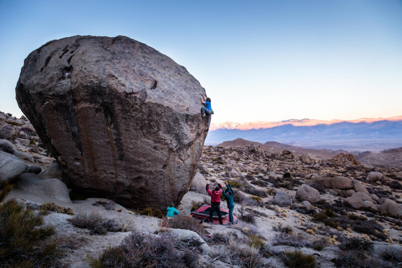 Climbers scaling a large boulder in a rocky, mountainous area at sunrise, with others watching below on crash pads. visit bishop