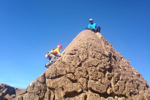 Child climbing a rocky peak with an adult seated at the top; both under a clear blue sky. visit bishop