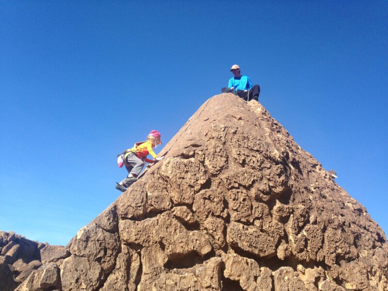 Child climbing a rocky peak with an adult seated at the top; both under a clear blue sky. visit bishop