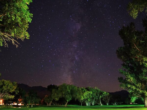 A nighttime scene of the Milky Way stretching across a star-filled sky above a dark landscape in Bishop, California, with illuminated green trees and a distant glowing building. visit bishop