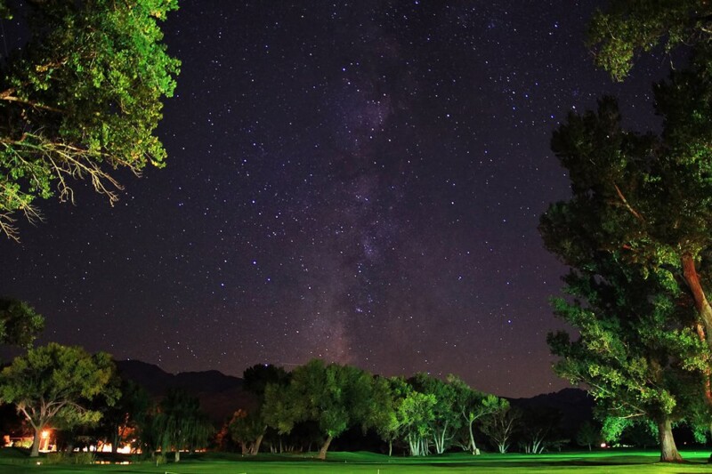 A nighttime scene of the Milky Way stretching across a star-filled sky above a dark landscape in Bishop, California, with illuminated green trees and a distant glowing building. visit bishop