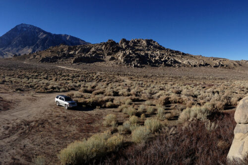 Panoramic view of a dry, rocky landscape with a white car parked in the middle. Mountains are visible in the background. visit bishop