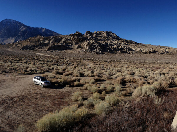 Panoramic view of a dry, rocky landscape with a white car parked in the middle. Mountains are visible in the background. visit bishop