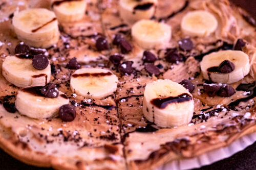 A close-up of a dessert pizza from Bishop, California, topped with banana slices, chocolate chips, and drizzled with chocolate sauce. The crust appears thin and crispy, with traces of powdered sugar sprinkled over the delectable toppings. visit bishop