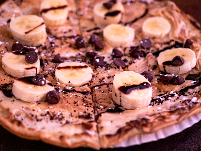 A close-up of a dessert pizza from Bishop, California, topped with banana slices, chocolate chips, and drizzled with chocolate sauce. The crust appears thin and crispy, with traces of powdered sugar sprinkled over the delectable toppings. visit bishop