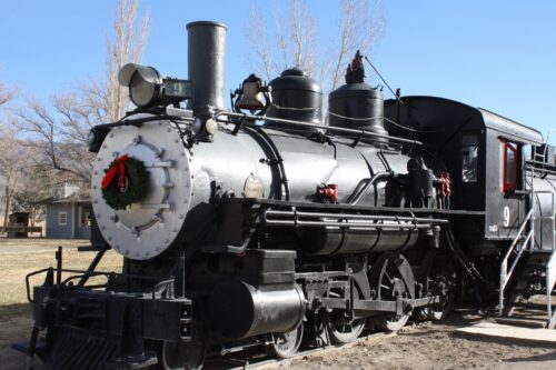 A vintage steam locomotive decorated with a wreath on its front, parked outdoors on a sunny day. visit bishop