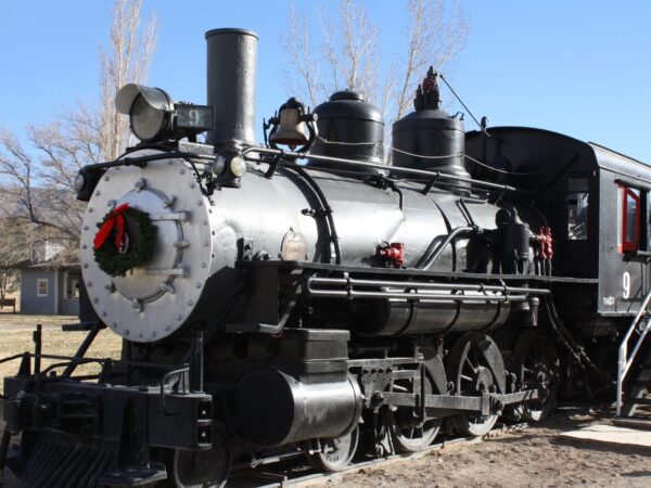 A vintage steam locomotive decorated with a wreath on its front, parked outdoors on a sunny day. visit bishop