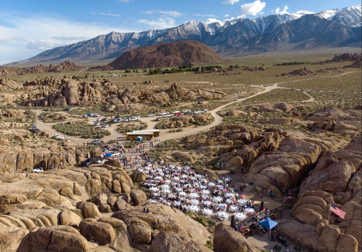 Aerial view of a large outdoor event with tables set among the rocky terrain, mountains, and blue sky in the background, capturing the stunning beauty of Bishop, California in the Eastern Sierra. visit bishop