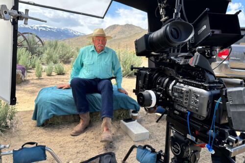 A man in a blue shirt and hat sitting on a cot outdoors with a professional film camera set up in front of him. visit bishop