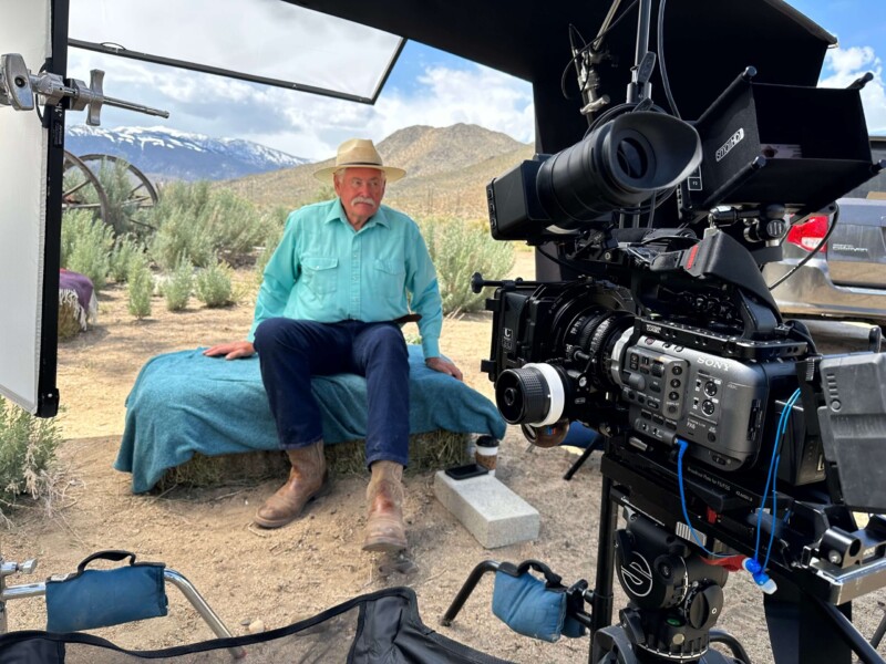 A man in a blue shirt and hat sitting on a cot outdoors with a professional film camera set up in front of him. visit bishop