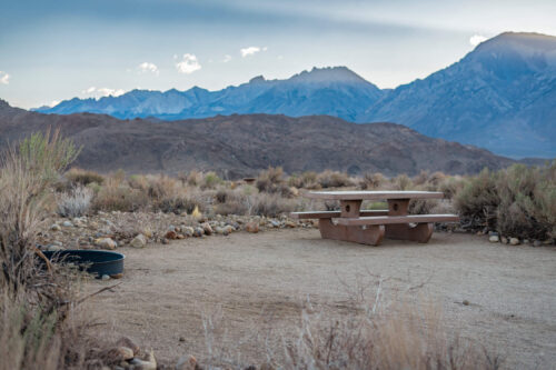 A solitary picnic table and fire ring are set in a dry, brush-covered landscape near Bishop, California, with a backdrop of rugged, towering mountains under a mostly clear sky with some clouds. The terrain appears arid and rocky, indicative of a high desert or mountainous region. visit bishop