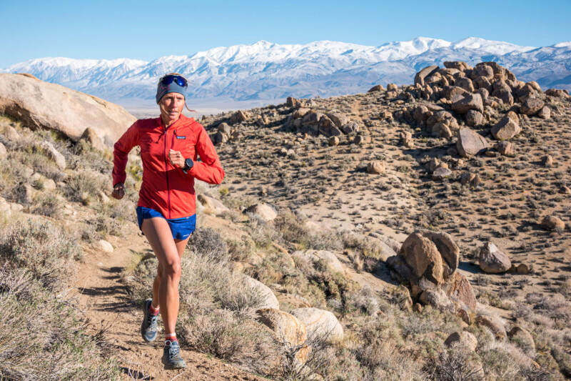 A person in a red jacket and blue shorts is running on a rocky trail through the desert landscape near Bishop, California, with large boulders around. Snow-covered mountains tower in the background under a clear blue sky. visit bishop