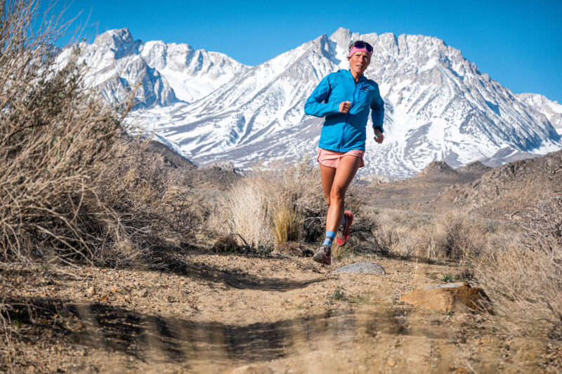 A person runs on a rugged trail in the mountainous area of Bishop, California. They are wearing a blue jacket, pink shorts, and sunglasses. Snow-capped peaks and clear blue skies form the breathtaking background landscape. Sparse vegetation lines the rocky trail. visit bishop