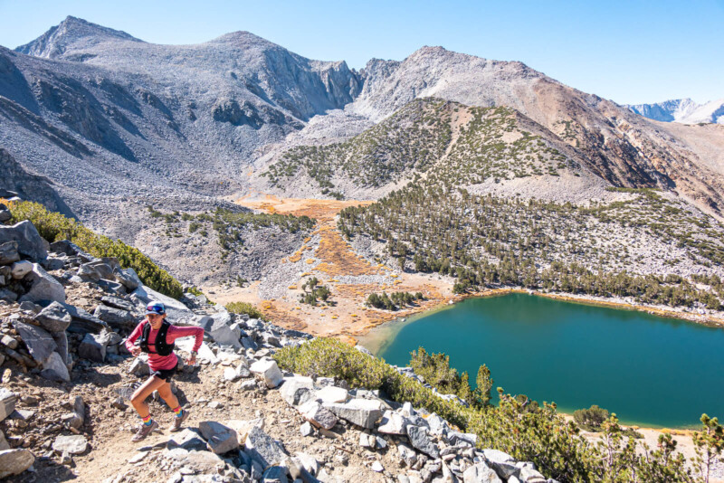 A hiker runs along a rocky trail overlooking a turquoise lake surrounded by mountains and sparse greenery. visit bishop