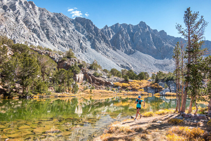 A person in athletic gear runs along the shore of a clear mountain lake, surrounded by trees and rocky peaks. visit bishop