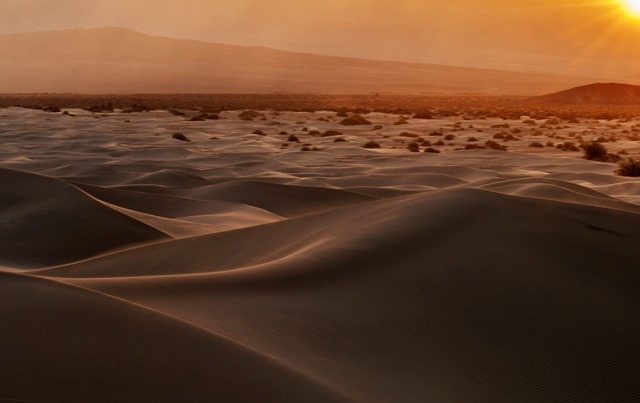 Golden sun setting over an expansive desert with gently rolling sand dunes and distant mountains. visit bishop