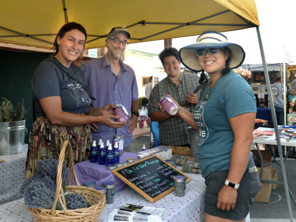 People smiling at a market stall in Bishop, California displaying lavender products, including a chalkboard sign, basket, and spray bottles. visit bishop