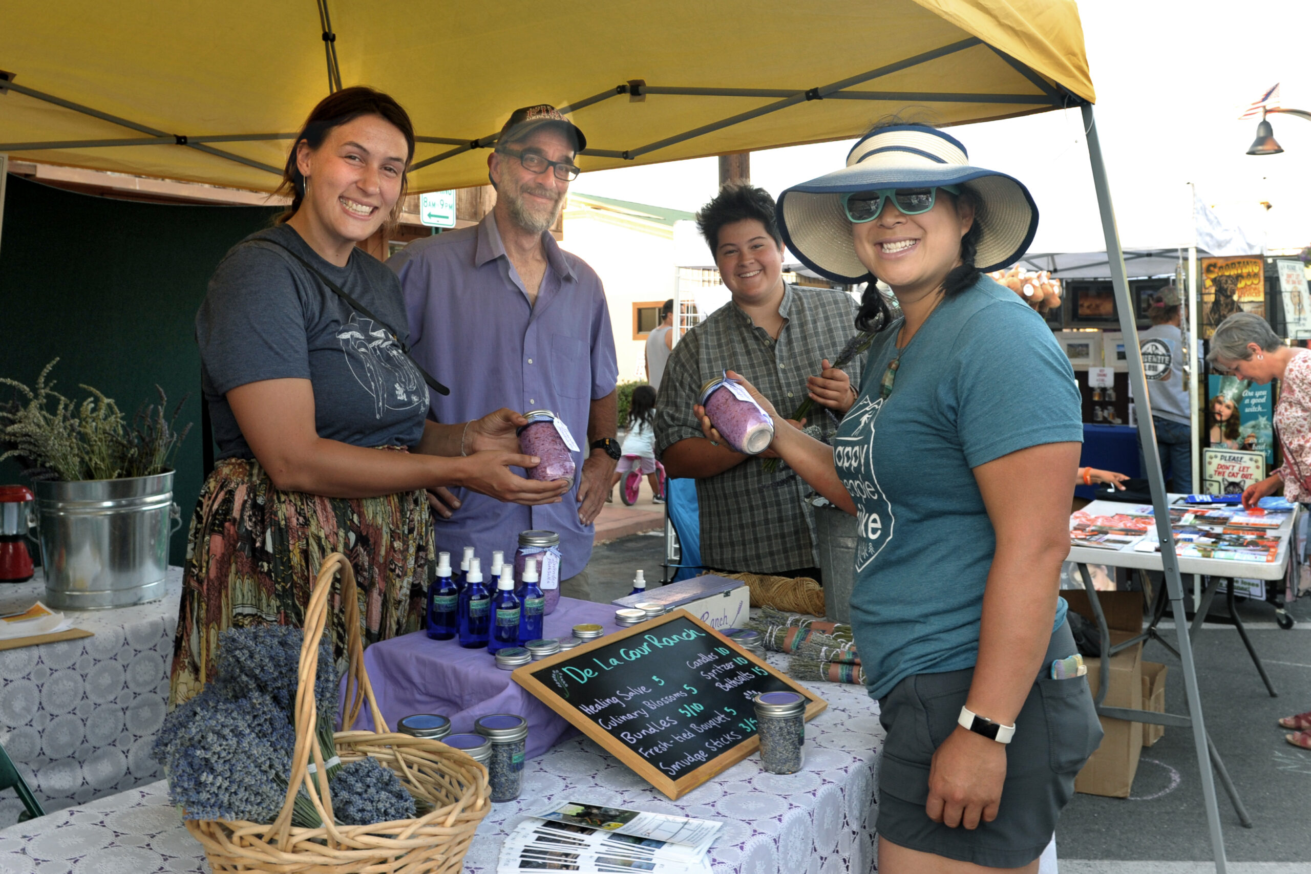 People smiling at a market stall in Bishop, California displaying lavender products, including a chalkboard sign, basket, and spray bottles. visit bishop