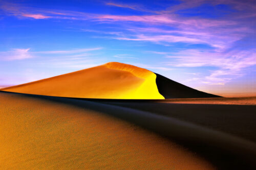 A large sand dune bathed in golden sunlight against a vibrant sunset sky with wispy clouds. visit bishop