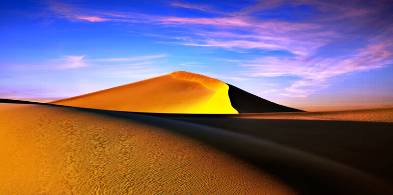 A large sand dune bathed in golden sunlight against a vibrant sunset sky with wispy clouds. visit bishop