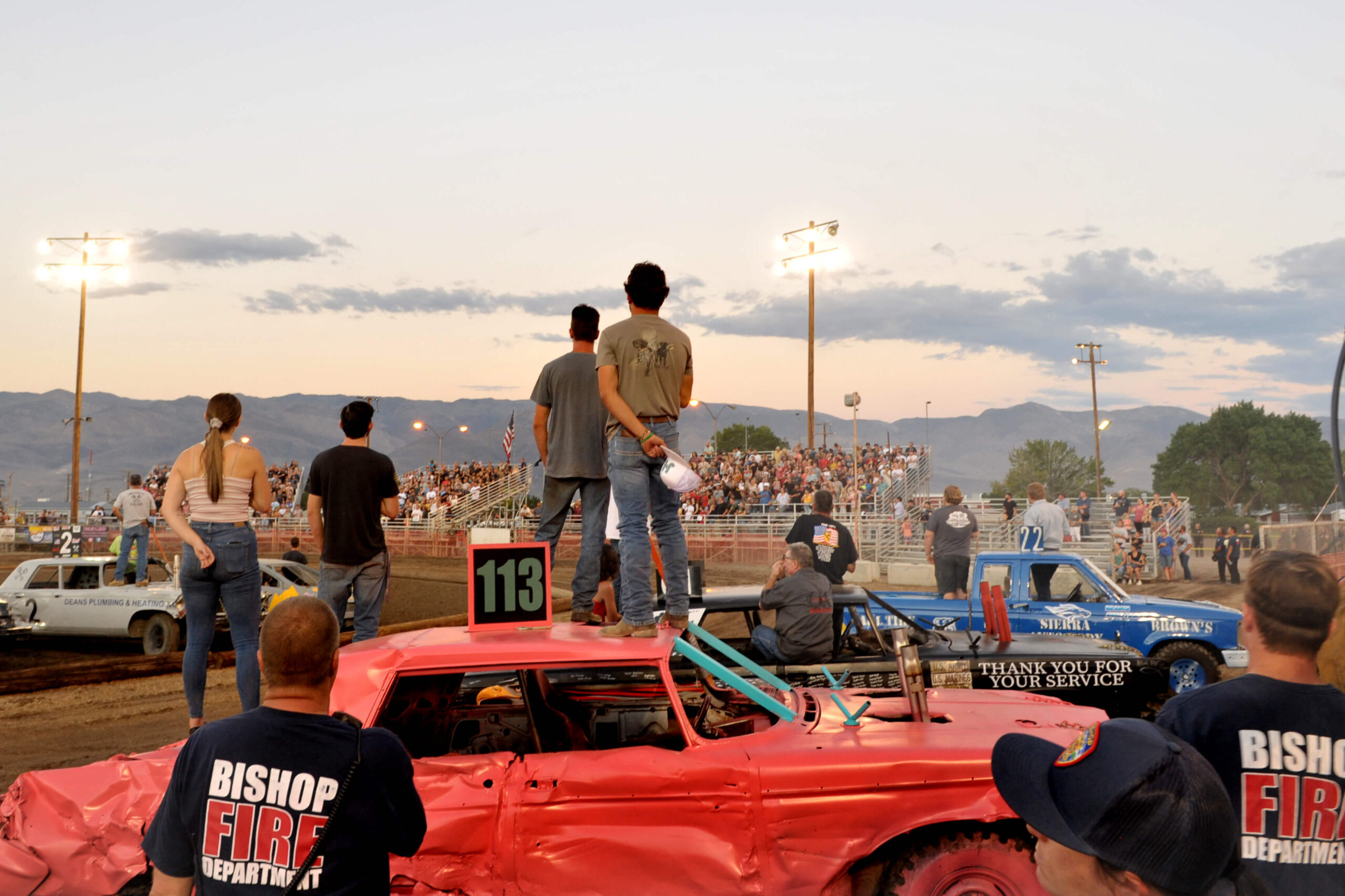 People at a demolition derby in Bishop, California, a person standing on a car marked "113," crowd watching, and the majestic Eastern Sierra mountains in the background. visit bishop