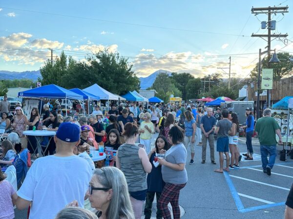 People enjoying an outdoor market in Bishop, California, with tents, vendors, and food under a bright sky with clouds and the Eastern Sierra mountains in the background. visit bishop