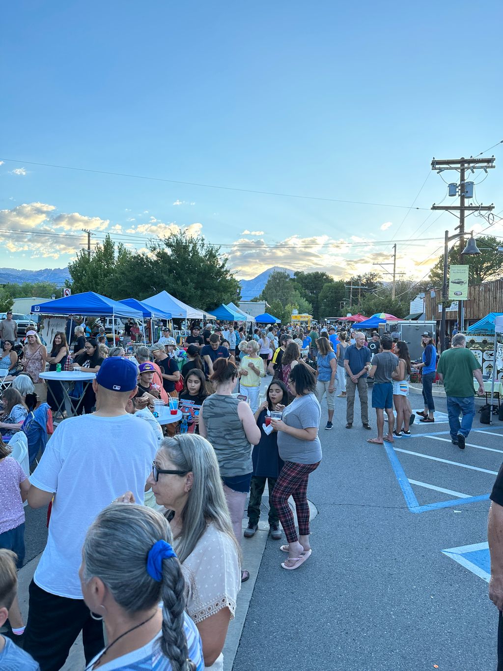 People enjoying an outdoor market in Bishop, California, with tents, vendors, and food under a bright sky with clouds and the Eastern Sierra mountains in the background. visit bishop