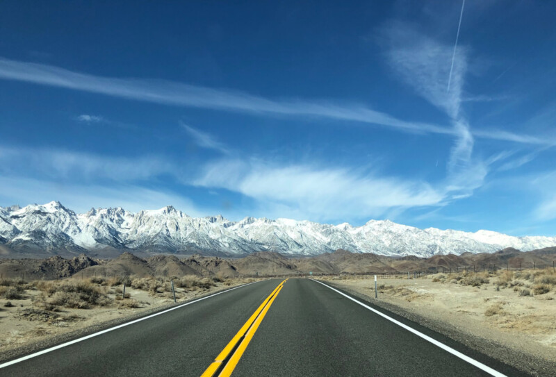 A straight road in Bishop, California, leads towards a snowy mountain range under a clear blue sky with wispy clouds. visit bishop