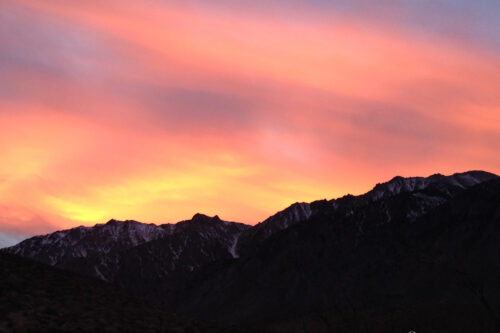 A vibrant sunset sky with shades of pink, orange, and yellow over a silhouette of a mountainous landscape. visit bishop