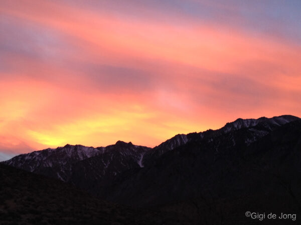 A vibrant sunset sky with shades of pink, orange, and yellow over a silhouette of a mountainous landscape. visit bishop