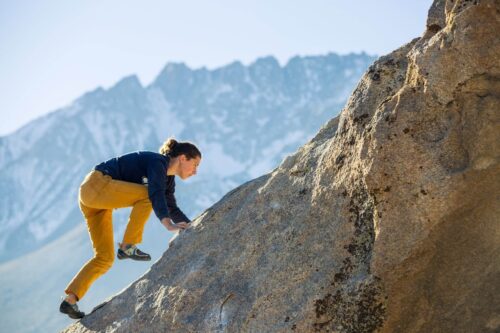 Person in yellow pants and blue shirt climbs a steep rock face with mountains in the background. visit bishop