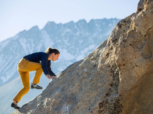 Person in yellow pants and blue shirt climbs a steep rock face with mountains in the background. visit bishop