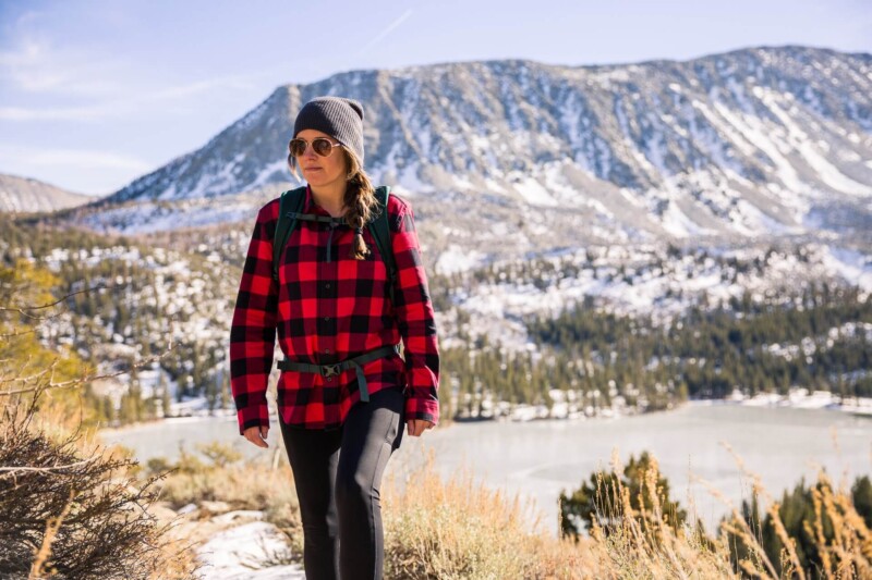 A woman in a red plaid shirt and beanie hikes in a snowy mountainous area, with a lake and mountains in the background. visit bishop