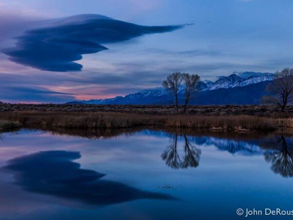 Two trees and snow-capped mountains reflecting in a calm lake under a dramatic, cloud-filled twilight sky. visit bishop