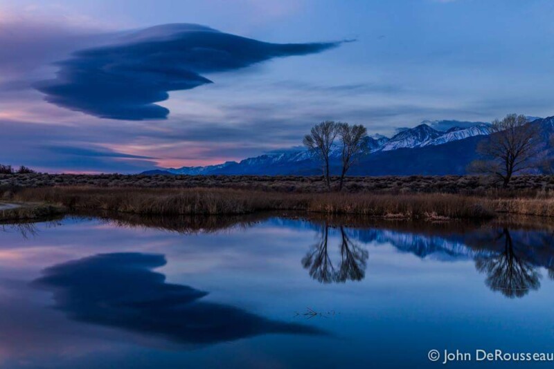 Two trees and snow-capped mountains reflecting in a calm lake under a dramatic, cloud-filled twilight sky. visit bishop