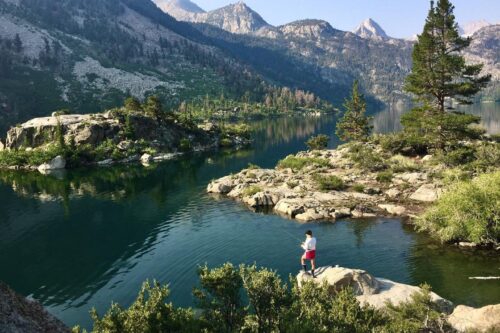 A person in a white shirt and red shorts stands on a rocky ledge overlooking a serene mountain lake near Bishop, California, surrounded by trees and rugged terrain. The lake is calm, reflecting the surrounding mountains with a clear, blue sky overhead. visit bishop