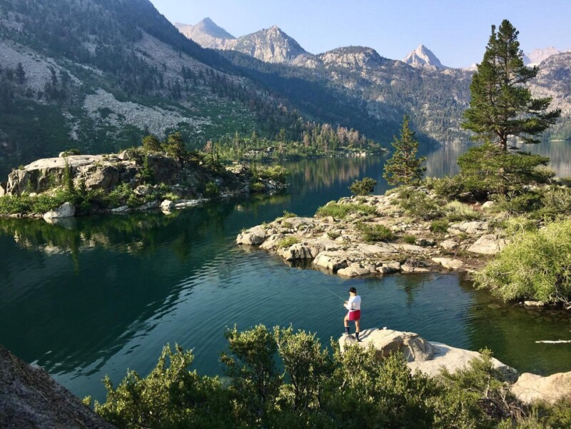 A person in a white shirt and red shorts stands on a rocky ledge overlooking a serene mountain lake near Bishop, California, surrounded by trees and rugged terrain. The lake is calm, reflecting the surrounding mountains with a clear, blue sky overhead. visit bishop