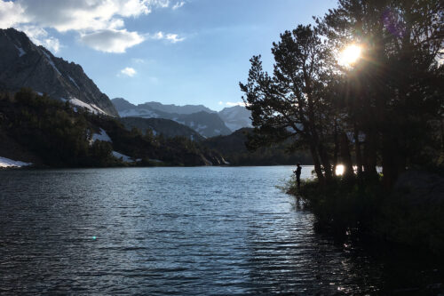 A serene lake scene with surrounding mountains, trees, and a person silhoutted against the sun. (©Gigi de Jong). visit bishop