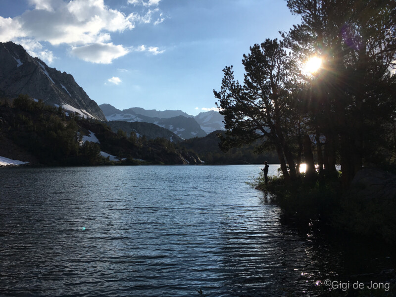 A serene lake scene with surrounding mountains, trees, and a person silhoutted against the sun. (©Gigi de Jong). visit bishop