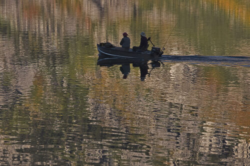 Two people fish on a small boat, gliding over a calm, reflective water surface with autumn colors in the background. visit bishop