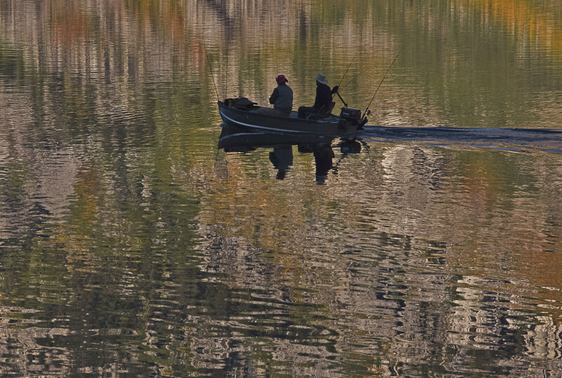 Two people fish on a small boat, gliding over a calm, reflective water surface with autumn colors in the background. visit bishop