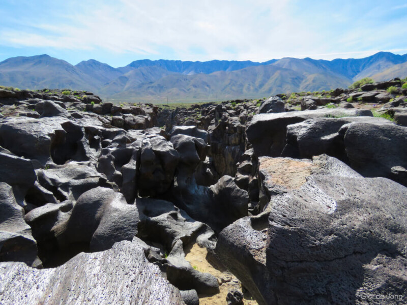 Rocky terrain with large cracks and crevices, set against a backdrop of mountain range under a bright, clear sky. visit bishop