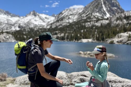 Two people wearing hiking gear sit on rocks by a lake with snow-capped mountains in the background. visit bishop